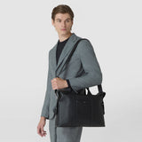 Front view: A man carries the SOFT BRIEFCASE IN CACHEMIRE LEATHER AND MOSAICO in black leather with woven decorative bands and a shoulder strap; “SERAPIAN” is displayed on the front.