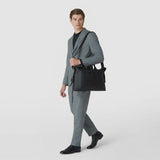 Front view: A man carries the SOFT BRIEFCASE IN CACHEMIRE LEATHER AND MOSAICO over his shoulder; the spacious black leather bag features textured Mosaico strips and short handles.
