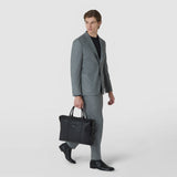 Front view: A man carries the SOFT BRIEFCASE IN CACHEMIRE LEATHER AND MOSAICO by its handles; the black leather bag with woven Mosaico details complements his gray suit.