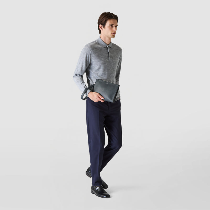 Frontal view: A person holds the CLUTCH IN CACHEMIRE LEATHER AND MOSAICO in navy blue leather with a wrist handle loosely in the right hand, elegantly dressed.