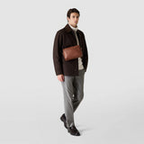 Frontal view: EAST WEST CROSSBODY BAG IN CACHEMIRE LEATHER shown worn across the chest by a man, highlighting its rectangular tobacco-brown leather design and oval silhouette.