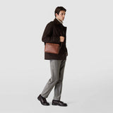 Frontal view: EAST WEST CROSSBODY BAG IN CACHEMIRE LEATHER shown worn crossbody by a person, featuring a compact oval tobacco brown leather bag with a subtle texture.