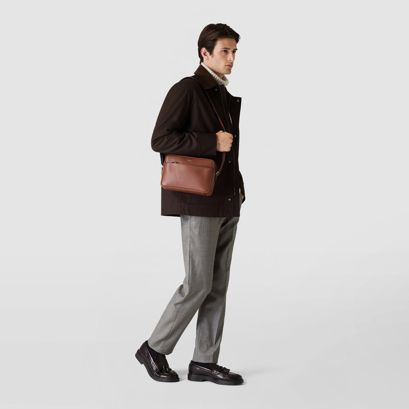 Frontal view: EAST WEST CROSSBODY BAG IN CACHEMIRE LEATHER shown worn crossbody by a person, featuring a compact oval tobacco brown leather bag with a subtle texture.
