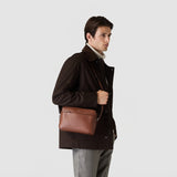 Frontal view: EAST WEST CROSSBODY BAG IN CACHEMIRE LEATHER worn by a person over the shoulder, showing a rectangular, tobacco brown leather bag with zip pocket.