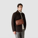 Frontal view: EAST WEST CROSSBODY BAG IN CACHEMIRE LEATHER worn crossbody by a man, showcasing its oval silhouette and smooth, tobacco-colored calfskin leather.