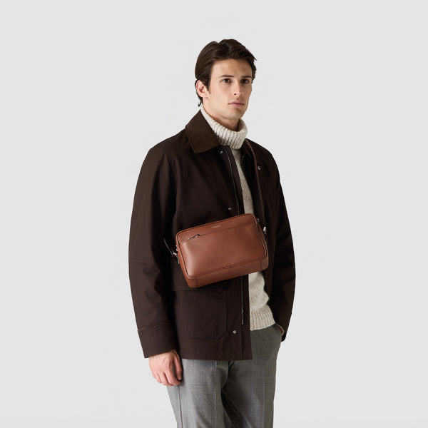 Frontal view: EAST WEST CROSSBODY BAG IN CACHEMIRE LEATHER worn crossbody by a man, showcasing its oval silhouette and smooth, tobacco-colored calfskin leather.