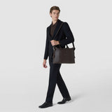 Front view: A man carries the SOFT BRIEFCASE IN CACHEMIRE LEATHER AND MOSAICO over his shoulder; the spacious espresso-colored leather bag features woven Mosaico details and short handles.