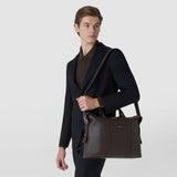 Front view: A man carries the SOFT BRIEFCASE IN CACHEMIRE LEATHER AND MOSAICO over his shoulder; the espresso-colored leather bag features wide handles, a zipper, and decorative Mosaico stripes.