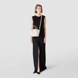 正面図: MOSAICO MICROCHAIN製ミニSECRETバッグ、小さな装飾チェーン付き。クリーム色のミニバッグが長い黒いドレスを着た女性の肩に掛けられ、無地の背景の前にある。