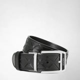 Detail view: The BELT IN MOSAICO is rolled up, showing black leather with a geometric pattern and a silver-tone buckle bearing the inscription “SERAPIAN” against a light background.