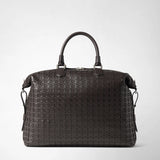 Front view: BRIEFCASE IN MOSAICO in dark nappa leather with a geometric woven pattern, two short handles, and a detachable shoulder strap stands against a light, neutral background.