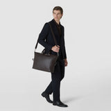 Front view: A man carries the BRIEFCASE IN MOSAICO over his shoulder; the spacious dark brown leather bag features an intricate geometric woven pattern against a light background.