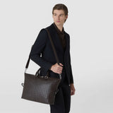 BRIEFCASE IN MOSAICO: Angled view of a dark brown leather bag with a geometric woven pattern and shoulder strap, worn by a man in a dark suit against a light background.