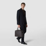 Front view: A man carries the BRIEFCASE IN MOSAICO by its handles; the dark brown leather bag features a woven geometric pattern against a light studio background.