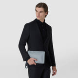 Front view: A man holds the CLUTCH IN MOSAICO in front of his black suit; the arctic blue leather bag features a geometric woven pattern, a zipper, and a wrist strap. The name “SERAPIAN” is printed on it.