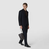 Front view: A man holds the CLUTCH IN MOSAICO by its wrist strap; the rectangular woven leather bag hangs beside his leg against a light studio background.
