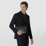Front view: A man holds the CLUTCH IN MOSAICO in front of his dark suit; the compact navy bag features woven leather, a zipper, and a wrist strap.