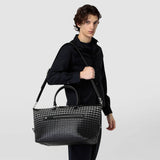 Front view: person carries TRAVEL BAG IN MOSAICO, a black-patterned travel bag in Mosaico nappa leather, casually over the shoulder with shoulder strap.