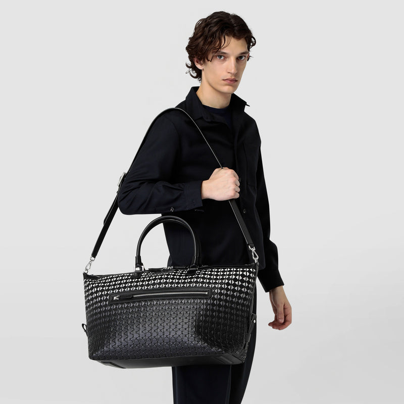 Front view: person carries TRAVEL BAG IN MOSAICO, a black-patterned travel bag in Mosaico nappa leather, casually over the shoulder with shoulder strap.