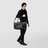 Front view: A person carries the TRAVEL BAG IN MOSAICO, a black-patterned, spacious travel bag in Chiaroscuro Mosaico nappa leather, over the shoulder.