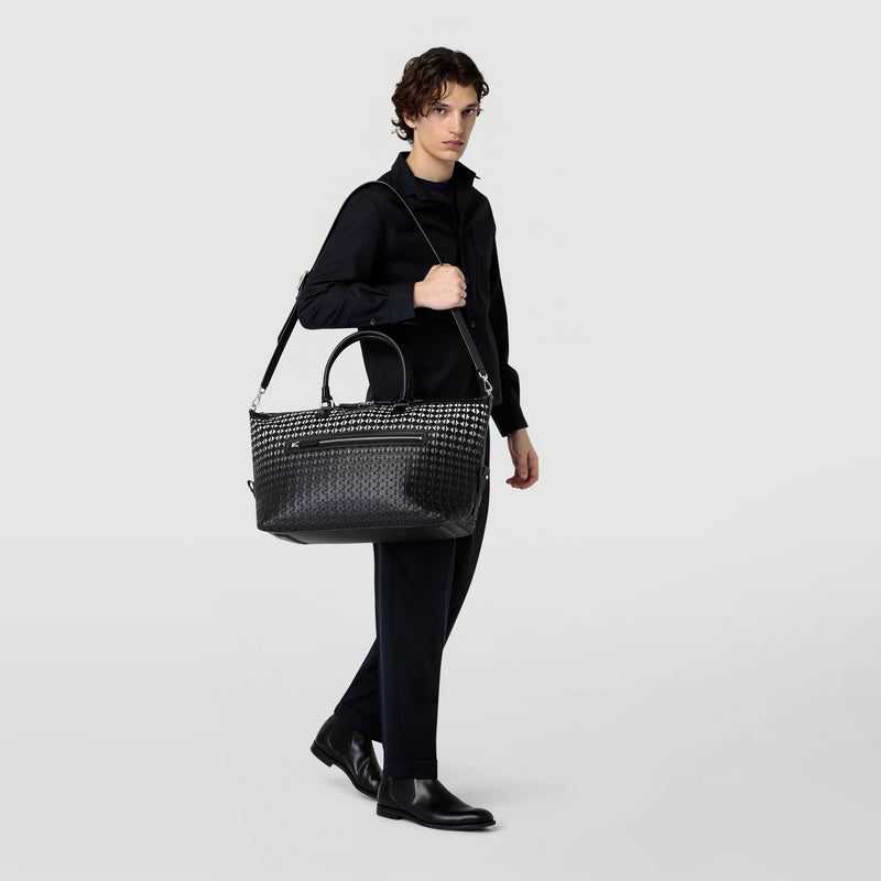 Front view: A person carries the TRAVEL BAG IN MOSAICO, a black-patterned, spacious travel bag in Chiaroscuro Mosaico nappa leather, over the shoulder.
