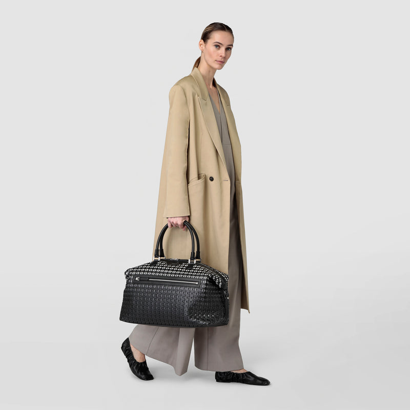 Front view of a woman in a beige coat holding the TRAVEL BAG IN MOSAICO made of Chiaroscuro Mosaico nappa leather with black and white weaving pattern.