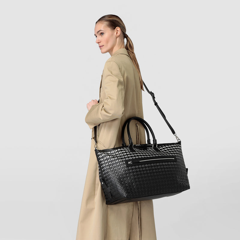 Side view: woman carrying the TRAVEL BAG IN MOSAICO, a large black travel bag made of patterned, woven Mosaico nappa leather, on a shoulder strap.