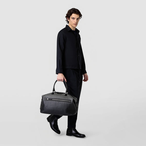 Front view of a man carrying the TRAVEL BAG IN MOSAICO in Chiaroscuro Mosaico nappa leather in black and white with woven pattern and two handles.