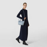 Front view: A woman wears the MINI SECRET BAG IN RIBBON MOSAICO with a slim shoulder strap; the icy blue, three-dimensional woven leather adds an elegant accent to her dark dress.