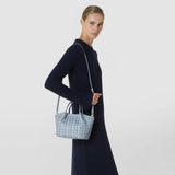 Front view: A woman carries the MINI SECRET BAG IN RIBBON MOSAICO by its shoulder strap; the ice-blue bag features a textured woven look and short handles against a light background.
