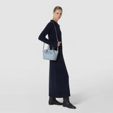 Front view: A woman wears the MINI SECRET BAG IN RIBBON MOSAICO with a narrow shoulder strap; the ice-blue bag features a textured geometric woven surface and short handles.