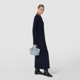 Front view: A woman carries the MINI SECRET BAG IN RIBBON MOSAICO in ice blue by the handle; the compact bag features a three-dimensional geometric woven pattern.
