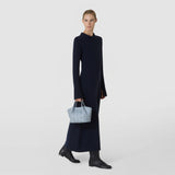 Front view: A woman carries the MINI SECRET BAG IN RIBBON MOSAICO in ice blue by its short handles, showing its intricate three-dimensional woven texture against a plain light background.