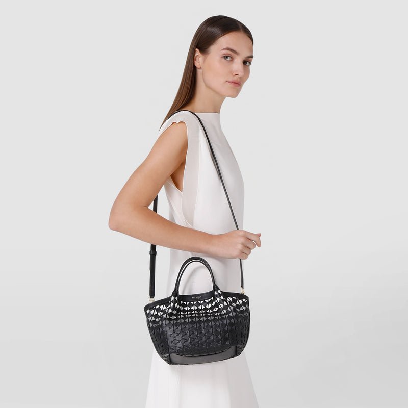 Side view: MINI SECRET BAG IN MOSAICO shown on a model wearing a white dress, featuring a compact, black woven nappa leather bag with short handles and a shoulder strap.