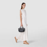 Side view: MINI SECRET BAG IN MOSAICO is held by a woman wearing a long white dress and gold sandals, showcasing the compact, woven leather bag in black.