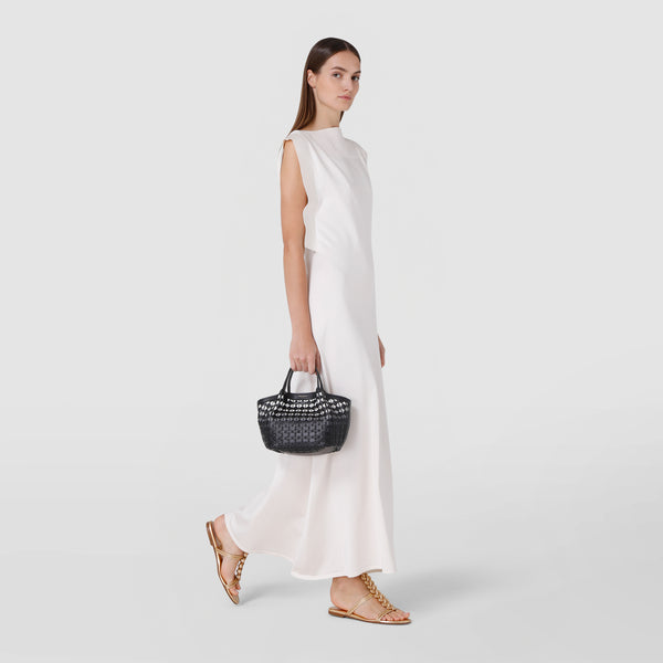 Side view: MINI SECRET BAG IN MOSAICO is held by a woman wearing a long white dress and gold sandals, showcasing the compact, woven leather bag in black.
