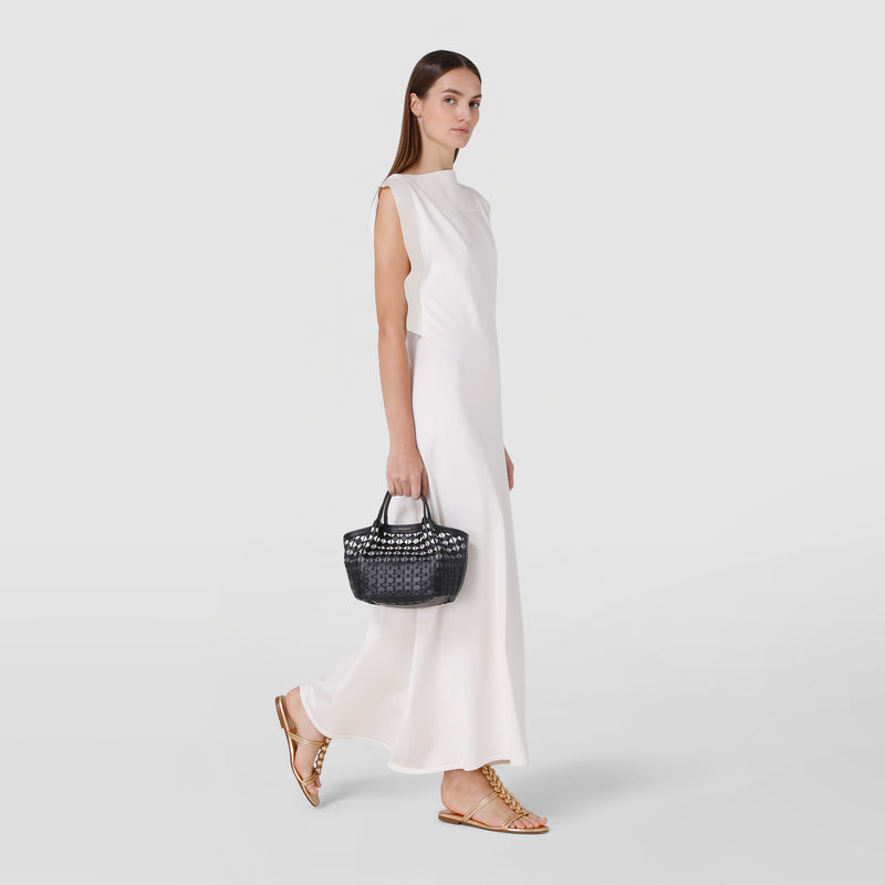 Side view: MINI SECRET BAG IN MOSAICO is held by a woman wearing a long white dress and gold sandals, showcasing the compact, woven leather bag in black.