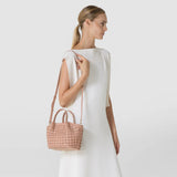 Front view: A woman carries the MINI SECRET BAG IN MOSAICO by its short handle and shoulder strap; the sandalwood-colored leather features a distinctive woven mosaic pattern.