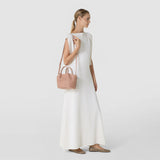 Front view: MINI SECRET BAG IN MOSAICO hangs from a woman's shoulder; the compact sandalwood-colored bag features a distinctive three-dimensional Mosaico weave and short handles.