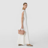 Front view: A woman carries the MINI SECRET BAG IN MOSAICO by its short handles. The compact sand-colored bag with an embossed mosaic pattern complements her white dress in a bright studio.
