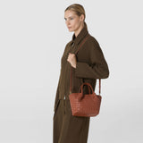 Front view: A woman carries the MINI SECRET BAG IN MOSAICO with a narrow shoulder strap; the brick-red leather bag features an intricate woven pattern and short handles.