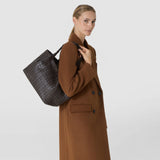 Front view: A woman carries the SMALL SECRET TOTE BAG IN RIBBON MOSAICO over her shoulder; the dark brown leather bag features a distinctive three-dimensional woven pattern.
