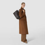 Side view: A woman carries the SMALL SECRET TOTE BAG IN RIBBON MOSAICO over her shoulder. The dark brown leather bag features a distinctive woven pattern and complements her long brown coat against a light background.