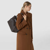 Side view: A woman carries the SMALL SECRET TOTE BAG IN RIBBON MOSAICO over her shoulder; the dark leather features a dimensional woven Mosaico pattern.