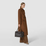 Front view: A woman carries the SMALL SECRET TOTE BAG IN RIBBON MOSAICO, made of dark, shiny Mosaico leather with two shoulder straps, against a light studio background.