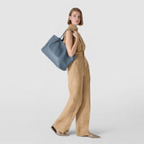 側面: 女性が肩にMOSAICO製SECRETトートバッグを掛けている; インディゴのMOSAICO編みナッパレザー製トートバッグ、大きく柔らかい、明るいニュートラルな背景の前に。