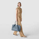 サイドビュー: 女性が歩き、MOSAICO製SECRETトートバッグ（インディゴ）を二本のハンドルでカジュアルに体の横に持っている、無地の薄いグレーのスタジオ背景の前で。