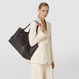 Front view: A woman carries the SECRET TOTE BAG IN RIBBON MOSAICO over her shoulder. The spacious dark brown leather bag features a distinctive woven mosaic pattern.