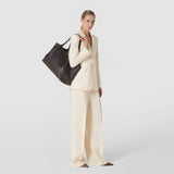 Front view: A woman carries the SECRET TOTE BAG IN RIBBON MOSAICO over her shoulder; the spacious ebony leather bag features a three-dimensional woven pattern.