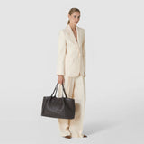 Front view: A woman carries the SECRET TOTE BAG IN RIBBON MOSAICO in dark nappa leather with a woven texture and long handles, paired with a light-colored outfit.